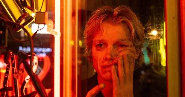 The image depicts a person in a dimly lit environment with red and yellow lighting, appearing distressed or contemplative.