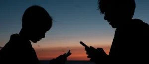Two individuals are silhouetted against a sunset sky, each engrossed in their smartphones.