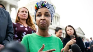 A woman in a green shirt and colorful headscarf speaks to reporters in front of a historic building.