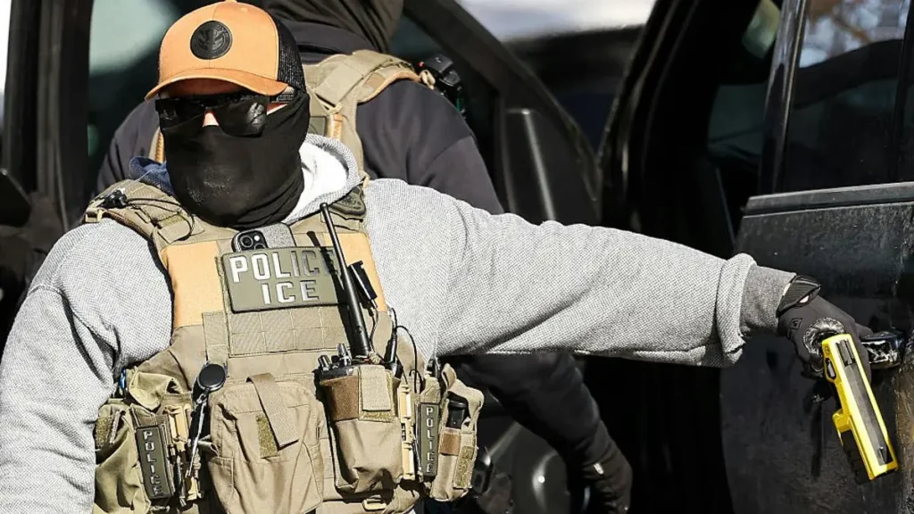 A law enforcement officer wearing tactical gear and a mask is seen outside a vehicle, holding a device in one hand.