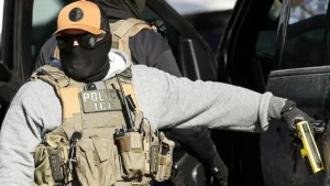 A law enforcement officer wearing tactical gear and a mask is seen outside a vehicle, holding a device in one hand.