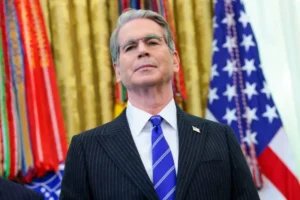 The image shows a man in a suit and tie standing in front of a backdrop featuring the American flag and other flags, suggesting a formal or official setting.
