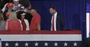 The image shows a political rally with attendees wearing patriotic attire and a man in a suit standing near a podium with an American flag design.