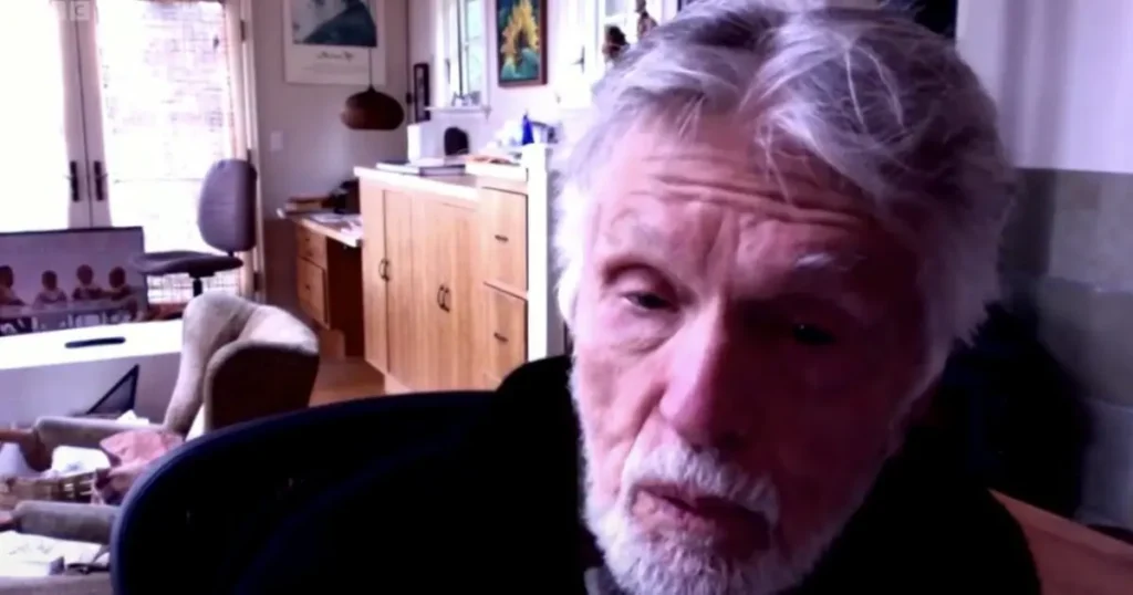 The image shows a person with gray hair and a beard, seated in a room with a wooden cabinet and various items in the background.