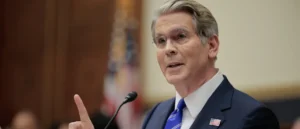 The image shows a man in a suit and tie speaking into a microphone, with an American flag in the background, suggesting a formal or governmental setting.