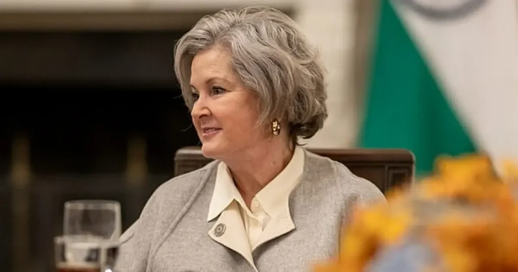 The image shows a woman in formal attire seated at a table, with a blurred background featuring a flag.