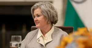 The image shows a woman in formal attire seated at a table, with a blurred background featuring a flag.