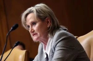 The image shows a woman speaking at a formal event, likely a press conference or a public meeting, as indicated by the microphones in front of her.