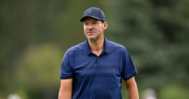 The image shows a man wearing a navy blue polo shirt and a matching cap, standing outdoors with a blurred green background.