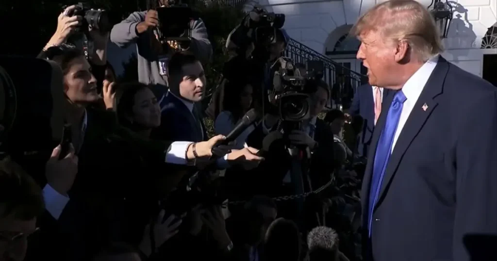 The image shows a public figure being interviewed by reporters and photographers in front of a building.