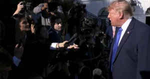 The image shows a public figure being interviewed by reporters and photographers in front of a building.