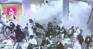 The image shows a group of people in a crowded area, possibly a public space, with some individuals appearing to be in distress or reacting to an incident.