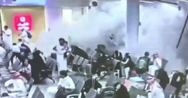 The image shows a group of people in a crowded area, possibly a public space, with some individuals appearing to be in distress or reacting to an incident.