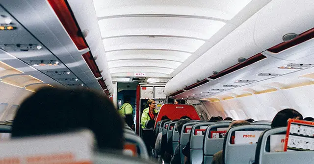 The image shows the interior of an airplane with passengers seated and a flight attendant walking down the aisle.