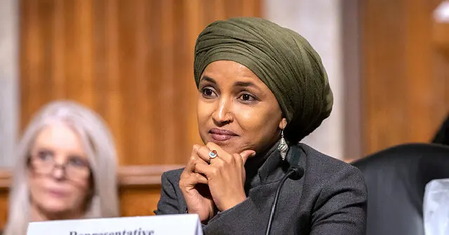 The image shows a person wearing a green turban and a suit, sitting at a table with a microphone, appearing to be engaged in a formal discussion or hearing.