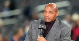 The image shows a man in a suit holding a microphone, likely providing commentary or analysis in a sports broadcast setting.