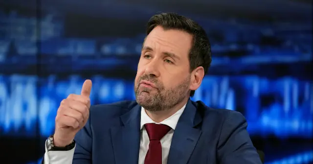 A man in a suit and tie gives a thumbs-up gesture in a news studio setting.