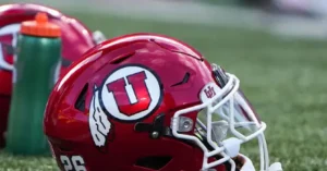 A close-up of a red football helmet with a "U" logo and a white face mask, resting on a field.