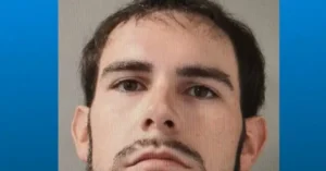 The image appears to be a mugshot of a man with short dark hair and a mustache, set against a blue and gray background.