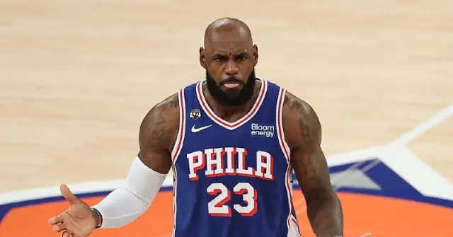 The image shows a basketball player wearing a Philadelphia 76ers jersey with the number 23, gesturing with his hand on the court.