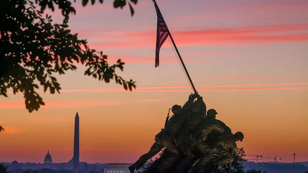 The iconic Iwo Jima Memorial stands prominently in the foreground against a backdrop of the Washington Monument and a vibrant sunset sky.