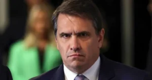 The image shows a man in a suit and tie, appearing serious and focused, with a blurred background suggesting a formal or professional setting.