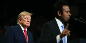 The image shows two individuals, one in a suit with a red tie and another in a suit with a blue tie, standing at a podium, likely during a political event or debate.