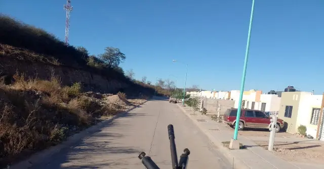The image shows a road in a rural or semi-urban area with a hillside on the left, a few buildings on the right, and a clear blue sky above.