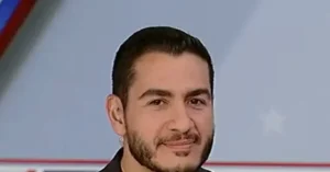 The image shows a person with short dark hair and a beard, wearing a dark suit, against a background featuring a red and white design.