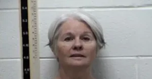 The image appears to be a mugshot of an individual with gray hair, standing against a wall with a height measurement scale on the left side.