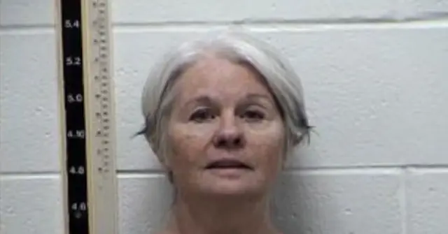 The image appears to be a mugshot of an individual with gray hair, standing against a wall with a height measurement scale on the left side.