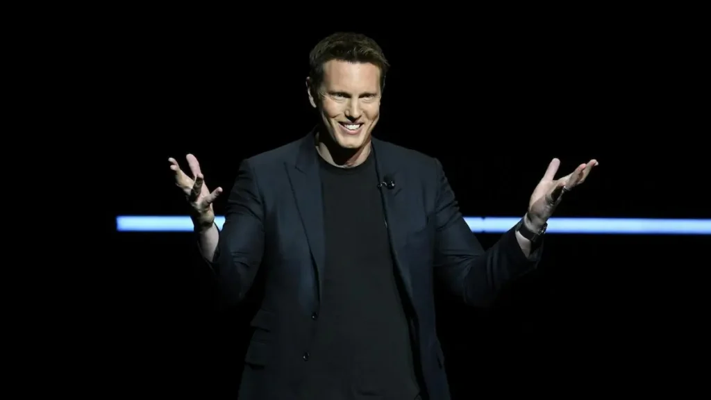 The image shows a person on stage, dressed in a dark suit, gesturing with their hands as they speak, with a microphone clipped to their lapel.
