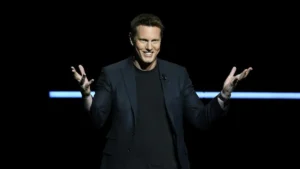 The image shows a person on stage, dressed in a dark suit, gesturing with their hands as they speak, with a microphone clipped to their lapel.