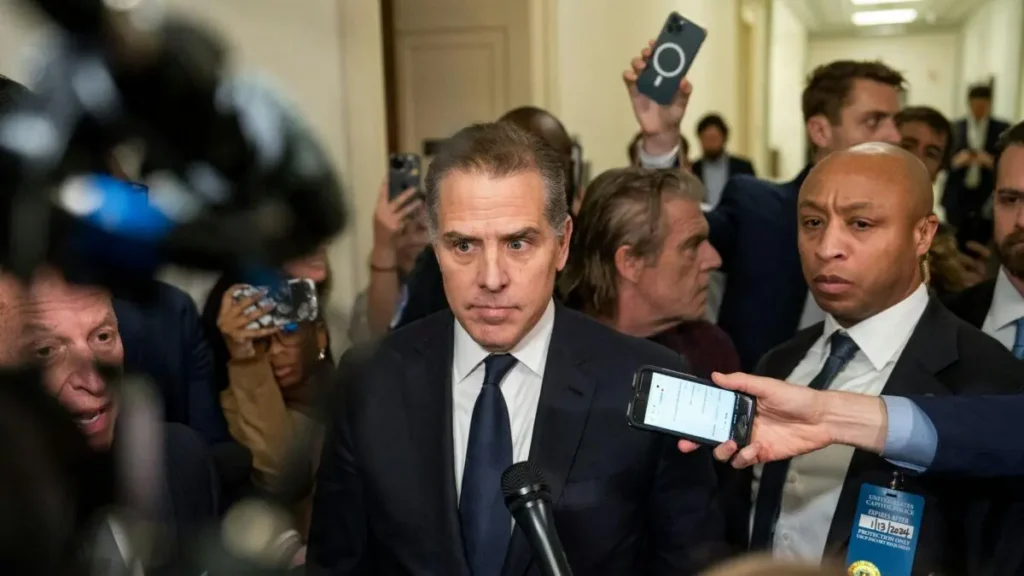 A man in a suit is surrounded by reporters and photographers as he speaks into a microphone.