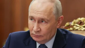 The image shows a person in formal attire, seated and looking to the side, with a serious expression.