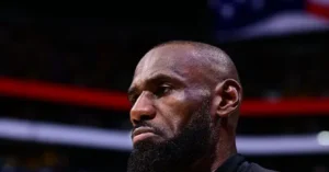 The image shows a close-up of a person with a shaved head and a beard, wearing a dark shirt, set against a blurred background that appears to be an indoor sports arena.
