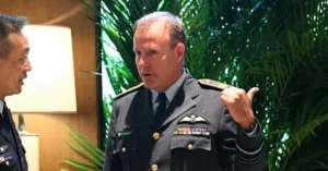 The image shows two individuals in military uniforms engaged in a conversation, with one gesturing while speaking.