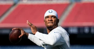 The image shows a football player in a white uniform and cap, preparing to throw a football on a field.