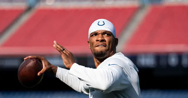 The image shows a football player in a white uniform and cap, preparing to throw a football on a field.