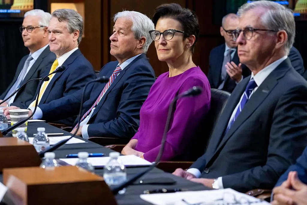 A group of individuals, including a woman in a purple top, are seated at a table with microphones, suggesting a formal meeting or hearing.