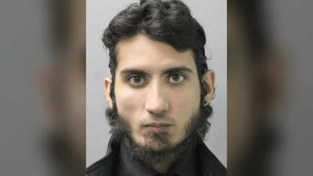 The image appears to be a mugshot of a person with dark hair and a beard, wearing a dark shirt.
