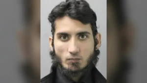 The image appears to be a mugshot of a person with dark hair and a beard, wearing a dark shirt.