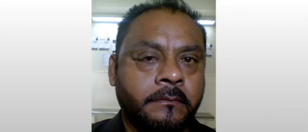 The image shows a man with a beard and mustache, wearing a dark shirt, in what appears to be an indoor setting with a light-colored wall and some papers or notices in the background.