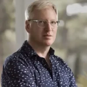 The image shows a person wearing a patterned shirt and glasses, standing in an outdoor setting with a blurred background.