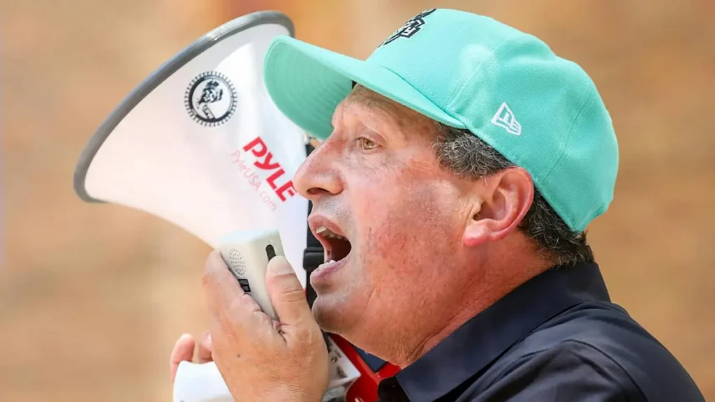 A man wearing a teal cap and a dark shirt is shouting into a megaphone.