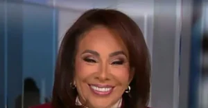 The image shows a person with shoulder-length brown hair, wearing a red top and gold earrings, smiling in front of a blurred background.