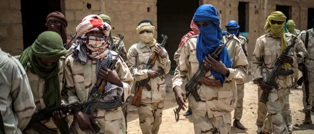 A group of armed individuals wearing camouflage uniforms and headscarves are seen holding weapons in a desert-like environment.