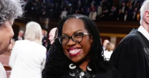A woman smiles warmly at a gathering, surrounded by other attendees in a formal setting.