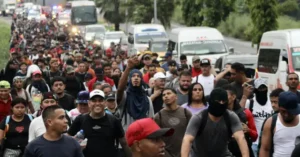 A large group of people is seen walking along a road, with some individuals taking photos and others appearing to be part of a migrant caravan.
