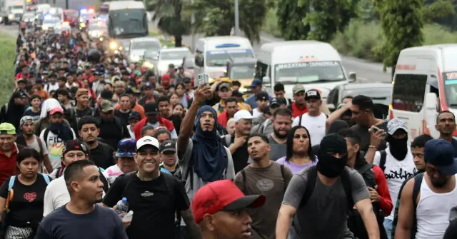 A large group of people is seen walking along a road, with some individuals taking photos and others appearing to be part of a migrant caravan.